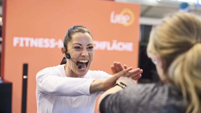 Auf der Fitness-Mitmachfläche der f.re.e 2025 motiviert eine Trainerin mit Headset eine Besucherin zu Sportübungen.