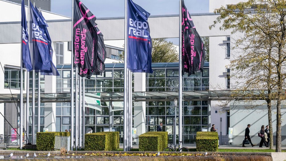 Expo-Real-Fahnen vor einem modernen Glasgebäude, mit vorbeilaufenden Menschen und Bäumen im Vordergrund.