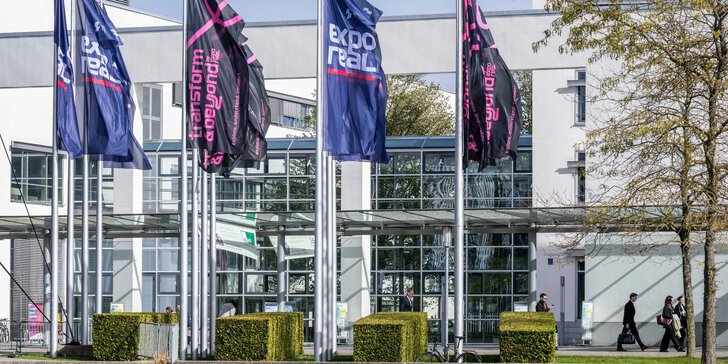 Expo-Real-Fahnen vor einem modernen Glasgebäude, mit vorbeilaufenden Menschen und Bäumen im Vordergrund.