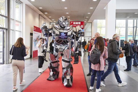 Ein großer silberner Roboter läuft auf einem roten Teppich durch einen Gang der transport logistic, umgeben von Menschen.