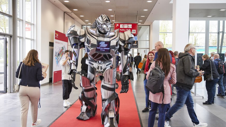 Ein großer silberner Roboter läuft auf einem roten Teppich durch einen Gang der transport logistic, umgeben von Menschen.