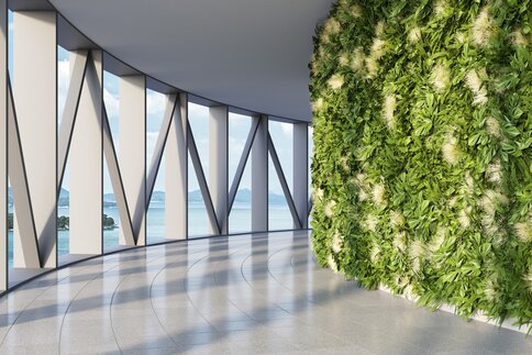 A modern, curved, bright hallway. The right wall is lushly planted with plants, the left side consists of triangular metal struts with a view of water and islands.