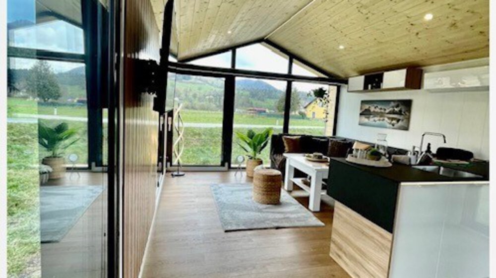 Modernes Tiny-House-Interieur mit Holzböden, großen Fenstern, Küche und Sitzbereich mit Blick auf eine malerische Landschaft.