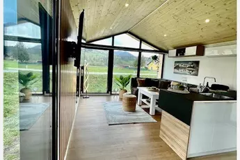 Modernes Tiny-House-Interieur mit Holzböden, großen Fenstern, Küche und Sitzbereich mit Blick auf eine malerische Landschaft.