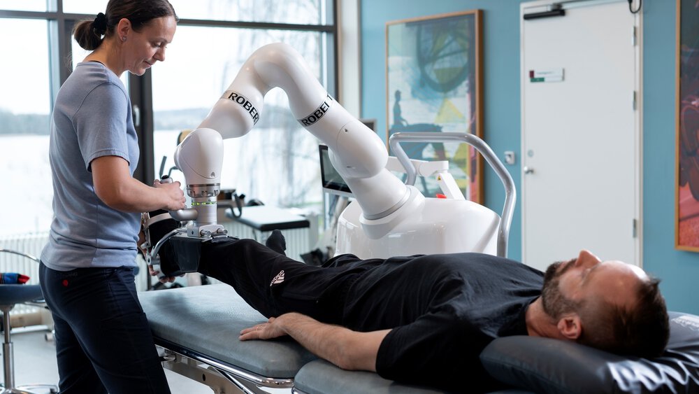 A therapist operates a robotic arm and helps a patient lying on a treatment table in a medical facility.