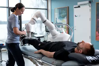 A therapist operates a robotic arm and helps a patient lying on a treatment table in a medical facility.