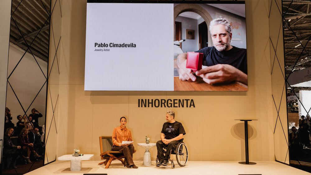 Two people are conversing on a stage beneath a large screen displaying a photograph and the name Pablo Cimadevila, jewellery artist.