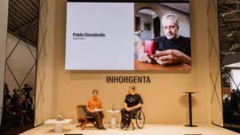 Two people are conversing on a stage beneath a large screen displaying a photograph and the name Pablo Cimadevila, jewellery artist.
