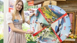 Eine lächelnde Frau im Dirndl steht an einem Stand auf der f.re.e Messe neben einem Glücksrad, welches nur Gewinne verspricht.