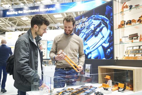 Two men examine components for electric vehicles at a trade fair stand.