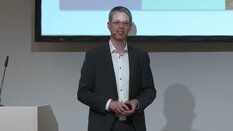 Ein Mann im Anzug und mit Brille spricht auf einer Bühne, ein Bildschirm steht dahinter.