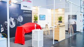 Moderner Messestand mit Produktgrafiken, Infowänden, Hochtischen und einem abgedeckten Exponat in einer Ausstellungshalle.