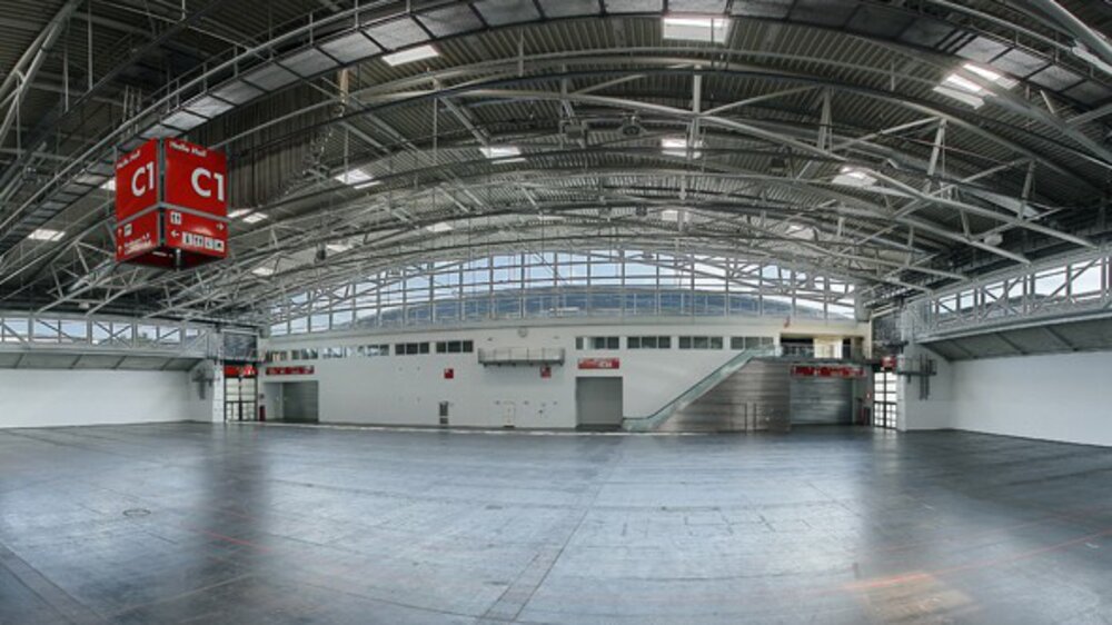 Geräumige, leere Industriehalle mit einem geschwungenen Metalldach, großen Fenstern und roten Schildern mit der Aufschrift „C1“, die von der Decke hängen.