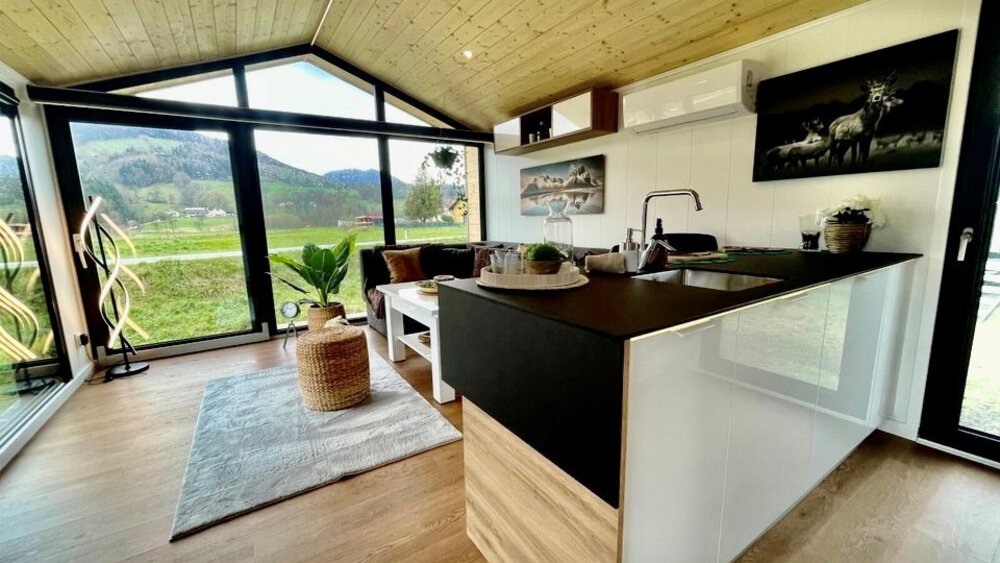 Modernes Interieur eines winzigen Hauses mit Küche, Wohnbereich, Holzdecke, großen Fenstern und malerischer Aussicht.