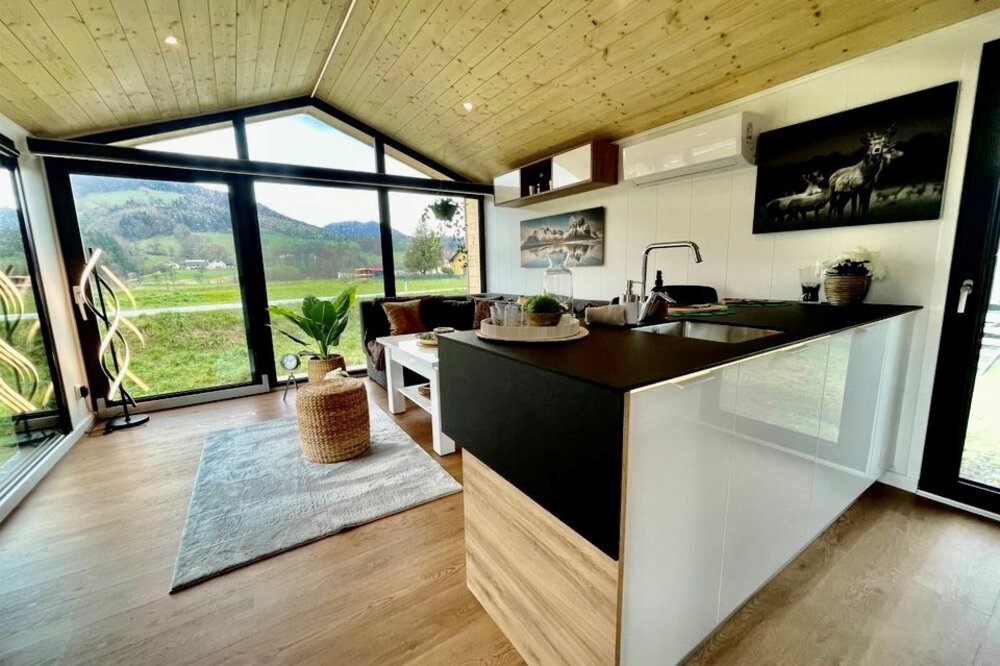 Modernes Interieur eines winzigen Hauses mit Küche, Wohnbereich, Holzdecke, großen Fenstern und malerischer Aussicht.