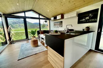 Modernes Interieur eines winzigen Hauses mit Küche, Wohnbereich, Holzdecke, großen Fenstern und malerischer Aussicht.