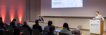Ein Redner spricht vor einem Publikum in einem Konferenzraum mit großer Leinwand und roter Beleuchtung an den Wänden