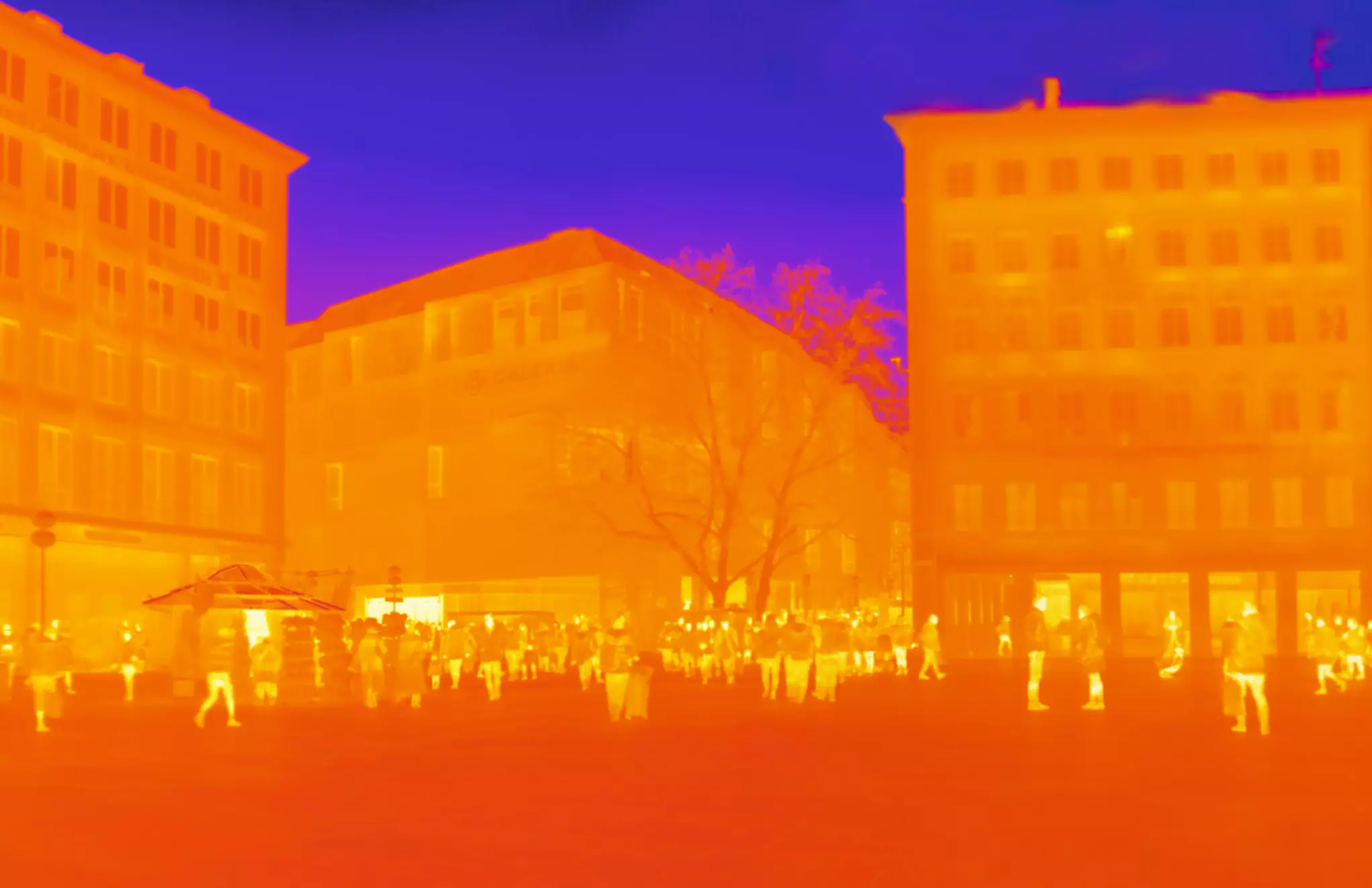 Thermal image of a city street with buildings and people in warm colours such as orange and red.