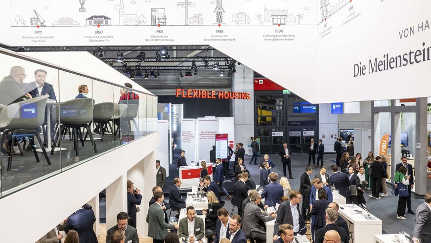 An exhibition booth at EXPO REAL in Munich with numerous visitors engaged in conversations or working at standing tables. On the upper level, several people are having business meetings in a modern lounge with glass railings. Above the booth, a timeline displays key milestones of the company’s history. In the background, other exhibition stands and a sign reading “Flexible Housing” can be seen.