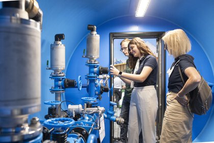 Three people are examining industrial equipment with pipes and valves in a tube-like room painted blue.