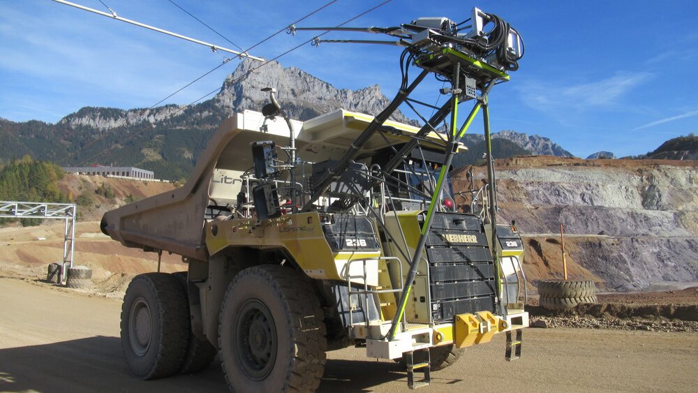 Großer Muldenkipper mit Oberleitung und Maschinen in einem Tagebau, Berge im Hintergrund.