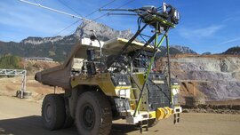 Großer Muldenkipper mit Oberleitung und Maschinen in einem Tagebau, Berge im Hintergrund.