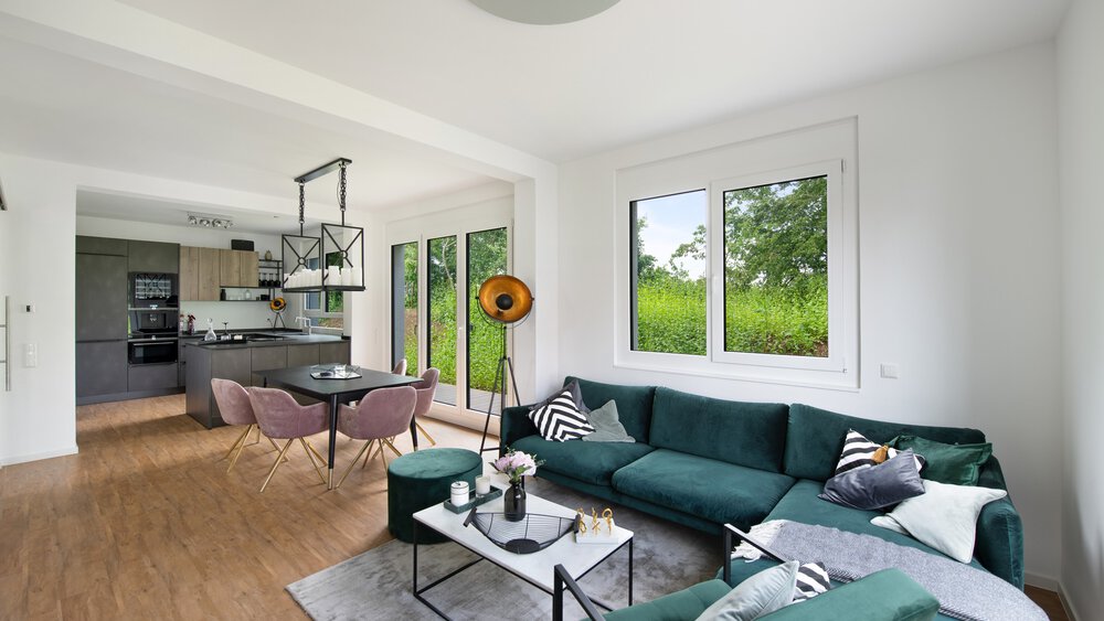 Modernes Wohnzimmer mit grünem Sofa, Couchtisch, Kissen, großen Fenstern und angrenzender Küche mit Essbereich.