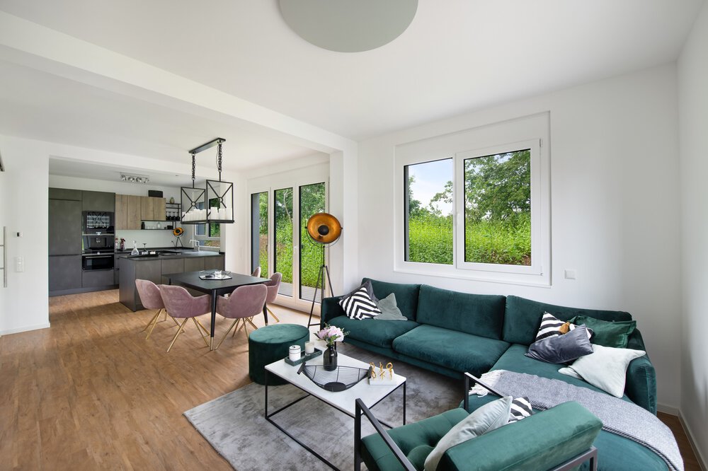 Modernes Wohnzimmer mit grünem Sofa, Couchtisch, Kissen, großen Fenstern und angrenzender Küche mit Essbereich.
