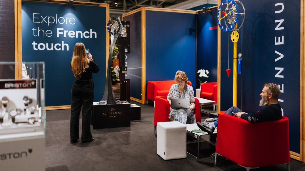 Ein Messestand auf der INHORGENTA: Eine Frau fotografiert eine Uhrskulptur, während zwei weitere lächelnd Personen in einer Sitzgruppe sitzen.