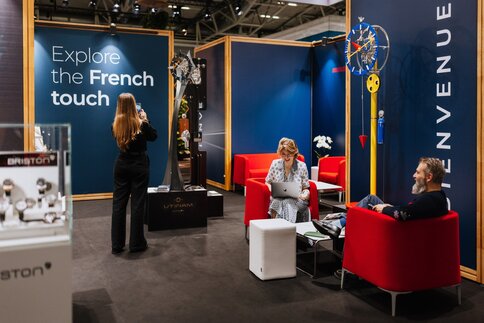 An exhibition stand at INHORGENTA: a woman is taking a photograph of a watch sculpture, whilst two other people are sitting in a seating area, smiling.