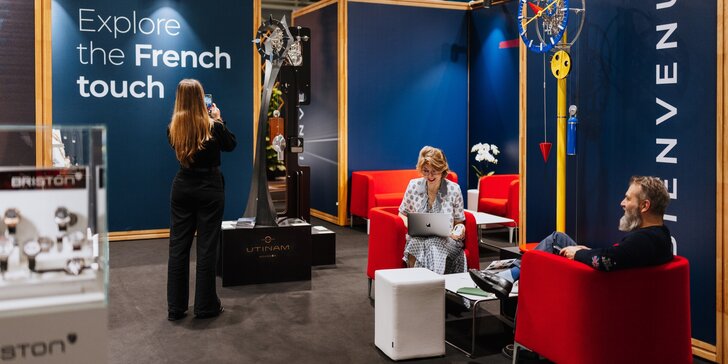 Ein Messestand auf der INHORGENTA: Eine Frau fotografiert eine Uhrskulptur, während zwei weitere lächelnd Personen in einer Sitzgruppe sitzen.