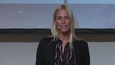 Eine blonde Frau spricht auf der Bühne, trägt ein Mikrofon-Headset und einen dunklen Blazer.