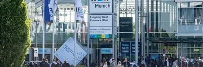 People walk to the entrance of IFAT Munich. Flags and part of the trade fair lake can be seen in the foreground.