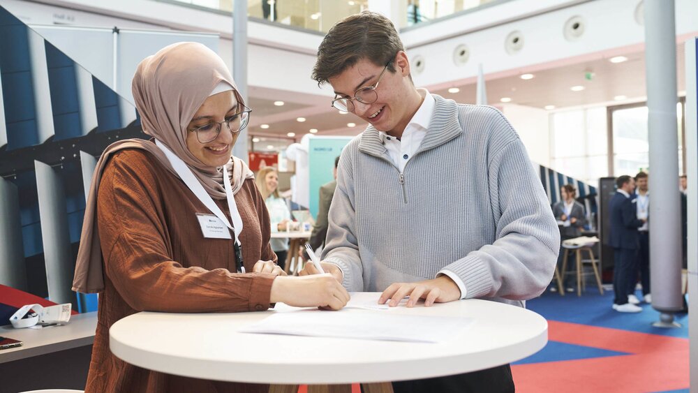 A young woman wearing a headscarf and a young man are signing a document together at a standing table during the EXPO REAL Career Day. Both are smiling as they fill out the form. In the background, other attendees of the career fair are seen engaged in conversations.