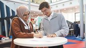 A young woman wearing a headscarf and a young man are signing a document together at a standing table during the EXPO REAL Career Day. Both are smiling as they fill out the form. In the background, other attendees of the career fair are seen engaged in conversations.