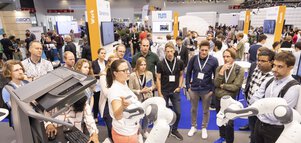 A group of people watch a woman demonstrating robots at a stand at a technology exhibition.