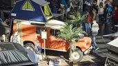 Ein orangefarbener Offroad-Wagen mit Dachzelt und Palmen-Deko auf einer Messe, umgeben von vielen Besuchern.