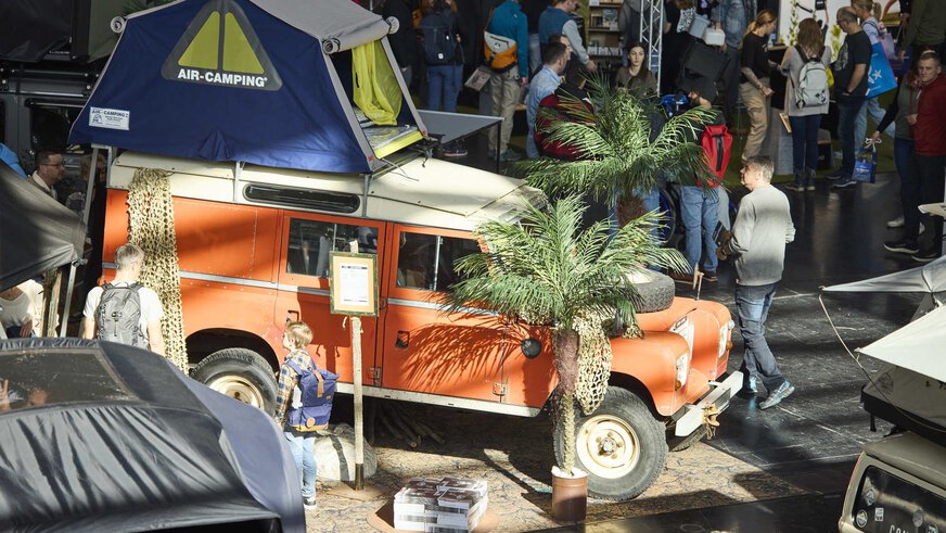 An orange off-road vehicle with a roof tent and palm tree decoration at a trade fair, surrounded by many visitors.