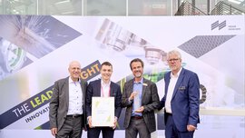 Vier Männer in Anzügen halten eine Auszeichnung und ein Zertifikat in den Händen. Im Hintergrund ist das Keyvisual der Laser zu sehen.