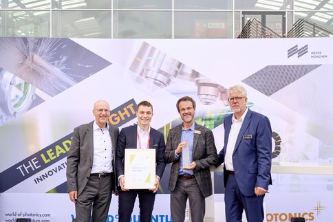 Four men in suits hold an award and a certificate in their hands. The key visual of the laser can be seen in the background.