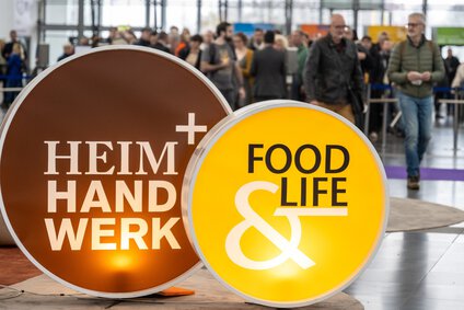 Auf dem Bild sind die Logos der Messen Heim und Handwerk, Food & Life im mit Menschen gefüllten Eingang der Messe München gezeigt.