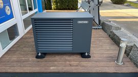 Eine graue Wärmepumpeneinheit steht auf einer Holzterrasse vor einem Gebäude in der Nähe eines Rasens und eines kleinen Metallpfostens.