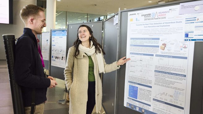 Eine Frau und ein Mann stehen vor einem Poster. Die Frau erklärt dem Mann etwas zu dem Poster. Beide lächeln