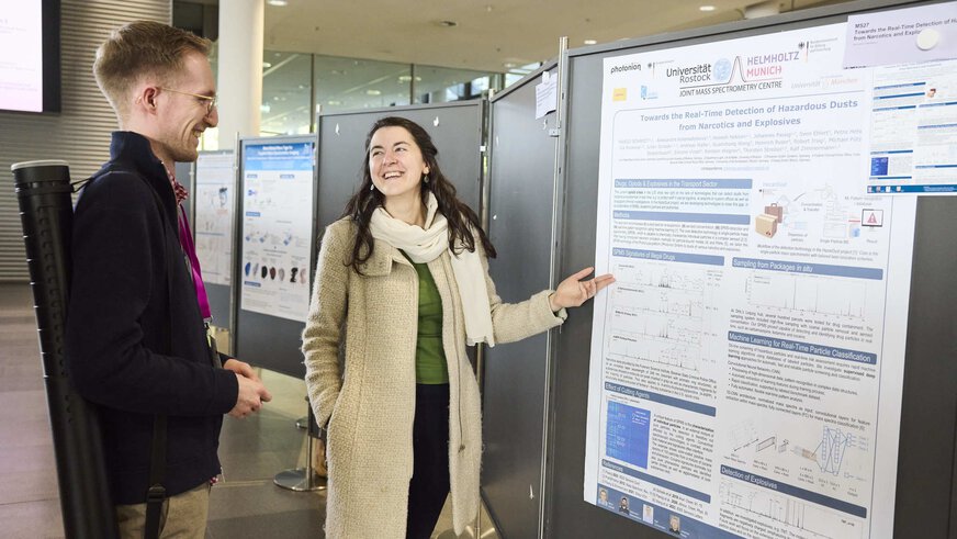 A woman and a man are standing in front of a poster. The woman explains something about the poster to the man. Both smile