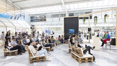 People sit on pallet sofas in front of a stage and listen to two speakers.