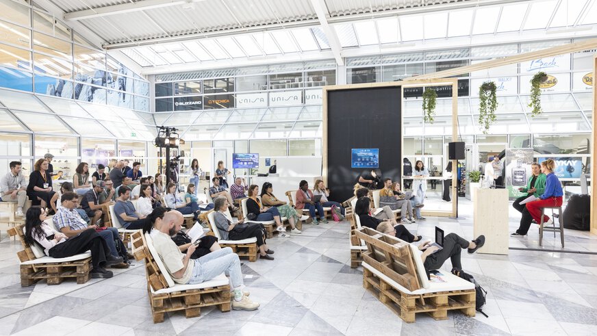 People sit on pallet sofas in front of a stage and listen to two speakers.