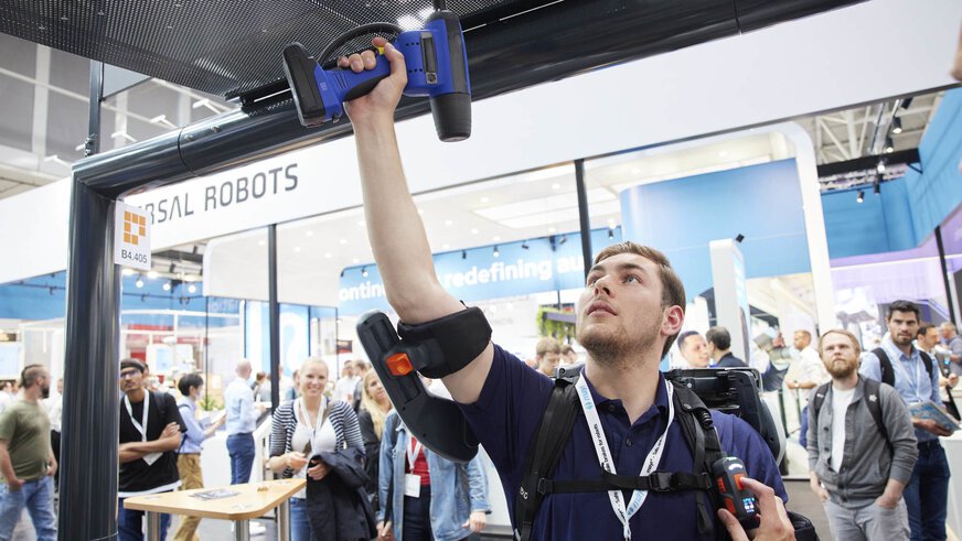 Person, die bei einer Robotik-Veranstaltung mit Exoskelett-Assistenz einen Akkuschrauber verwendet und von Zuschauern umgeben ist.