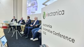 Five men are seated on a podium at the Electronica Automotive Conference 2025, with a conference sign in the foreground.