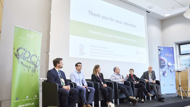 Eine Gruppe von fünf Personen sitzt vor einem Publikum auf einer Bühne mit Mikrofonen und Namensschildern und nehmen an einer Diskussion teil