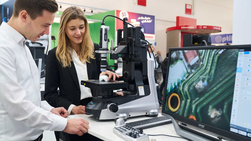 Ein Mann und eine Frau in Anzügen stehen an einem Mikroskop auf der productronica, während eine Kamera das Mikroskopbild auf ein Display überträgt.